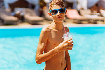 Portrait of a happy boy in sunglasses having a drink near the pool, child with a fresh drink in a luxury pool looking at the camera