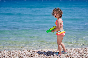 Happy cute little blonde girl is playing with water gun on the beach.         