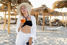 A Beautiful Blonde Woman On A Tropical Beach In A White Shirt Stands Under A Straw Umbrella Holds A Glass With An Alcoholic Cocktail In Her Hand And Enjoys.