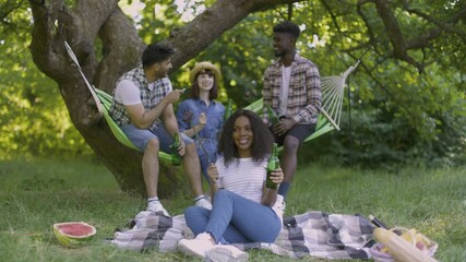 Wall Mural - Pretty african woman sitting on plaid at garden with bottle of beer and sausages in hands. Three diverse friends talking in hammock on background. Picnic time during summer season.