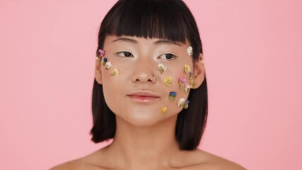 Wall Mural - A gorgeous asian woman posing to the camera with colorful flowers on her face standing over a pink background in the studio