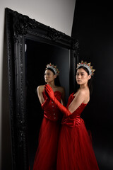 Wall Mural - Full length  portrait of beautiful young asian woman wearing red corset, long opera gloves and ornate crown headdress. Graceful posing against a full length mirror with a dark studio background.