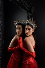 Wall Mural - Full length  portrait of beautiful young asian woman wearing red corset, long opera gloves and ornate crown headdress. Graceful posing against a full length mirror with a dark studio background.
