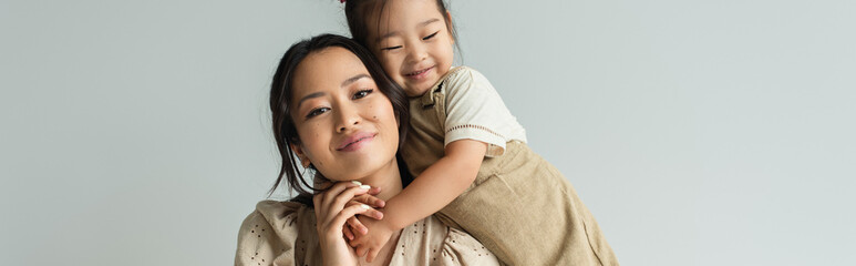 cheerful asian toddler daughter hugging happy mother isolated on gray, banner