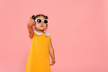 Canvas Print - asian toddler kid in yellow dress and sunglasses showing thumb up isolated on pink