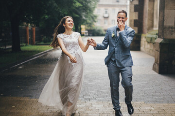 Wall Mural - Beautiful emotional wedding couple smiling and having fun in rain in european city. Provence wedding. Stylish happy bride and groom running on background of old church in rainy street.