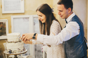 Wall Mural - Stylish happy bride and groom cutting together modern cake with lavender on background of festive table in stylish restaurant. Provence wedding reception. Beautiful wedding couple celebrating