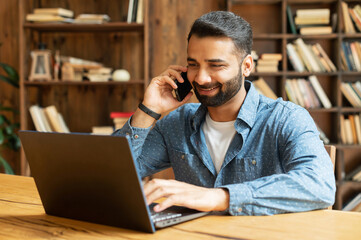 Wall Mural - Mid adult indian man working in his home office, using smartphone and laptop for communication with customers or colleagues, smiling multiracial eastern guy talking phone watching on computer screen