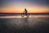Fototapeta Boho - Silhouette of a boy on a bicycle riding on a small portion of sand in the middle of the water from the baltic sea. Dramatic stunning sunset colors. Estonia, Europe