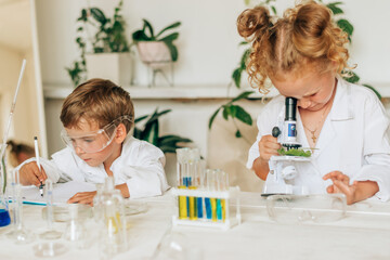 Wall Mural - Boy and girl in white uniforms and protective glasses do chemical experiments in a home laboratory.Back to school concept.Young scientists.Natural sciences.Preschool and school education of children.