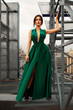 Attractive brunette girl in a long emerald dress posing on the catwalk on the roof against the background of the city, fashion concept, model in a luxurious dress against the background of urban