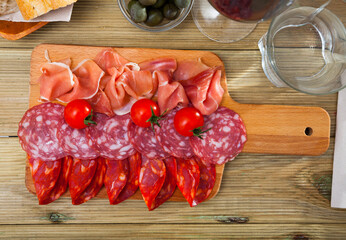 Top view of traditional Spanish meat platter - sliced dry-cured gammon and two sorts of sausages on wooden cutting board served with tomatoes, olives and wine..