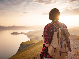 A young Asian woman with a backpack hiking in the summer and enjoying the view of the sunset sea and mountains. Mountain and coastal travel, freedom and an active lifestyle