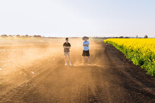 Two Boys Standing With Arms Crossed On Dusty Dirt Road Next To Canola Field On Farm