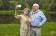 © Studio Romantic - Senior retired couple rest in green city park or forest. Happy mature woman standing making selfie portrait with husband on mobile phone camera. Old family having fun posing for photo recording video