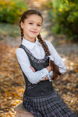 Wall Mural - Portrait of a cute schoolgirl in a school uniform sitting on a wooden chair among the autumn landscape