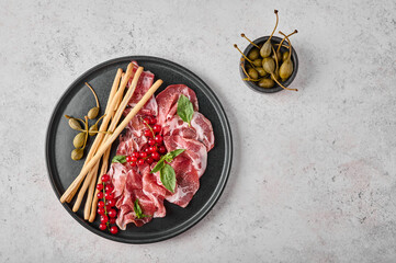 Slices of prosciutto coppa di parma ham with basil, grissini sticks, capers and redcurrant on dark plate on textured background. Top view with copy space