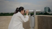 Young Traveler Woman Using Binoculars On Building Roof For Sightseeing And Urban Exploration. Adult Watching City Lifestyle With Telescope From Observation Point On Top Of Skyscraper
