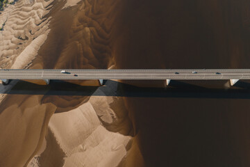 Wall Mural - Aerial drone shot of cars driving on bridge over river and sandy riverside at sunset. Amazing aesthetic top view shot with beautiful coast with dune sands. Drone aerial bridge with car traffic