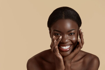 Wall Mural - Portrait close up of beautiful african girl. Joyful young woman touch her clean face and looking at camera. Concept of face skin care. Isolated on beige background. Studio shoot