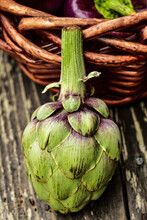 Closeup Of Single Green Artichoke With Purple Tinged Petals Leaning Against A Woven Basket On Rustic Wood Tabletop