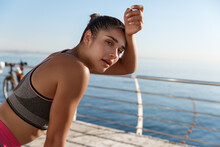 Close-up Of Attractive Sportswoman Taking A Breath During Workout, Looking At Camera And Wiping Sweat Off Forehead, Jogging Along Seaside Promenade