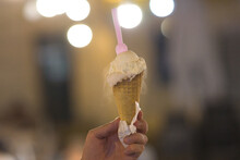 Hand Holding An Ice Cream Cone With A Pink Plastic Spoon In It On A Blurred Background At Night