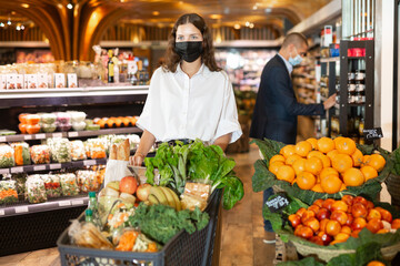 Woman in protective mask with shopping cart choosing groceries in supermarket