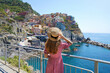 Traveler girl enjoying vacations in Italy. Young woman wearing hat and dress looking at italian village of Manarola with sea.