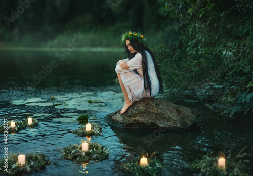 Slavic girl sits on stone on shore lake. Nymph fantasy woman hugs knees. Long black hair. Wreaths of grass, flowers float on water. Candles burning. River dusk forest green tree. Riutal of Divination