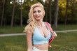 Portrait beautiful young blonde with sexy boobs in sporty blue fashionable top with pink stylish backpack at sunset in forest. Attractive pretty athletic girl model in sportswear for walk in park.