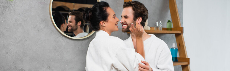 cheerful woman applying face cream on nose of smiling boyfriend in bathrobe, banner