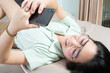 © Surachetsh - Beautiful glasses Asian woman lie on her back, chatting, typing, and texting, mobile in her bedroom with a pastel green - brown color theme.