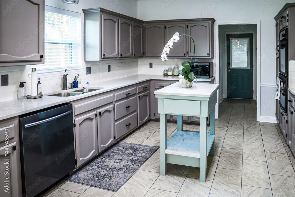 A renovated kitchen in an older home with painted gray cabinets, marble countertops, a small portable island and a tiled floor