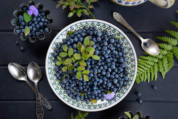 Wall Mural - Top view of plate with blueberries and spoons on dark wooden background, flat lay with fern leaf and blueberry leaves. Healthy design concept