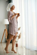 © Drazen - Full length of beautiful woman in nightgown drinks tea and looks through the window at home.