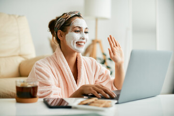 Wall Mural - Happy woman with hydrating facial mask waves during video call over laptop at home.