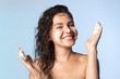 © deniskomarov - Portrait of a cheerful young woman washing face by lather having fun. Beautiful brunette with naked shoulders applying cleansing foam on facial skin on blue studio background. Skincare concept.