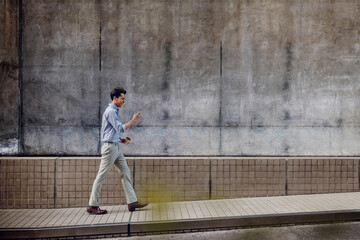 Smiling Young Asian Businessman in Casual wear Using Mobile Phone while Walking by the Urban Building Wall. Lifestyle of Modern People. Side View. Full Length