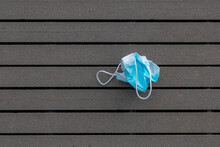Health Care And Protection From Virus Concept, A Piece Of Light Blue Surgical Face Mask Thrown Away On Black Wooden Floor After Use, Waste Procedure Or Medical Mask On Ground Of Street.