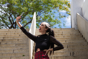 pretty young girl in punk style taking photos with her mobile phone on a staircase in the park. Technology and beauty concept