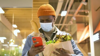 Wall Mural - Black man in disposable face mask walks along shopping mall supermarket holding products and typing on orange smartphone closeup