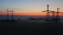 Aerial View Silhouettes High Voltage Steel Power Pylons In Field Covered With Fog Countryside. Misty Early Morning, Dawn. Drone Flight Low Over Power Transmission Lines. Electric Tower Line, Sunrise