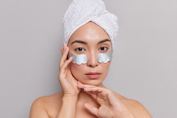 Wall Mural - Portrait of serious Asian woman keeps hands tenderly near face looks directly at camera enjoys softness of skin after spa procedures applies patches under eyes stands shirtless indoor grey background