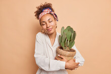 Indoor Shot Of Happy Dark Skinned Woman Gets Potted Cactus As Present Fond Of Houseplants Takes Care Of Succulent Plant Stands With Closed Eyes Wears Casual Shirt Kerchief On Head Beige Background