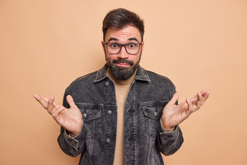 Wall Mural - Clueless hesitant bearded adult man spreads hands sideways in dismay feels indecisive has surprised unaware expression wears black denim jacket isolated over beige background. Human perception