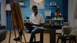 © DC Studio - Man of african american ethnicity designing sketch for vase drawing on white canvas and easel in workshop studio. Black artist with creativity and imagination creating art masterpiece