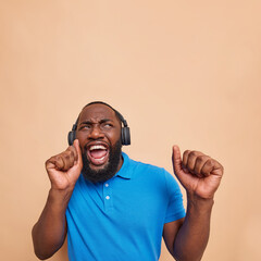 Wall Mural - Optimistic joyful dark skinned man dances carefree catches every bit of music has fun listens music via headphones raises arms wears casual blue t shirt isolated over beige background copy space