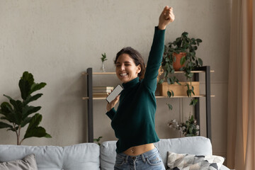 Wall Mural - Overjoyed young woman holding smartphone with white empty blank screen mockup, celebrating success, rejoicing online lottery win, received money refund or job promotion, standing in living room