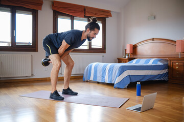 Bearded man doing kettlebell swing to workout at home with online class on laptop.